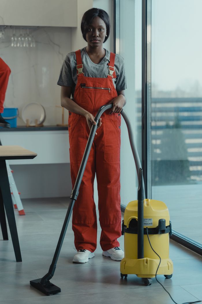 about-01 An adult woman in red coveralls operates a vacuum cleaner in a modern indoor setting, focusing on cleaning tasks.